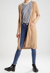Lång, beige cardigan i mjukt material; har öppen framsida, sidofickor och ribbade muddar, kombineras med randig topp och blå jeans.