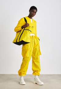 L'outfit impermeabile giallo presenta una maglietta dalla vestibilità ampia e pantaloni cargo con tasche. Una borsa a spalla gialla coordinata con dettagli neri completa il look.