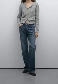 Gray cable-knit cardigan with a hood and gold buttons, paired with dark blue wide-leg jeans and black shoes. Simple, casual design.