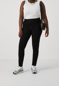 Jeans negros ajustados con cintura alta y cinco bolsillos, combinados con una camiseta sin mangas blanca y zapatillas plateadas. Incluye un bolso bandolera negro.