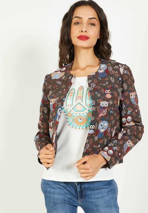 Mujer vistiendo una chaqueta marrón con un patrón de búhos coloridos sobre una camisa blanca que presenta un diseño de mano de Hamsa en azul y naranja.