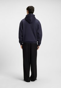 Uomo che indossa un felpa blu navy e pantaloni neri larghi, in piedi rivolto di spalle su uno sfondo grigio chiaro.