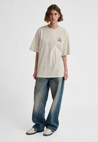 Béžové oversized tričko s krátkymi rukávmi, zdobené malým grafickým logom. V kombinácii s širokými vyblednutými modrými džínsami a bielymi teniskami.