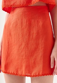 Mini gonna in lino arancione con superficie texturizzata e orlo scallop. Presenta una vita aderente e pieghe sottili per una forma accentuata.