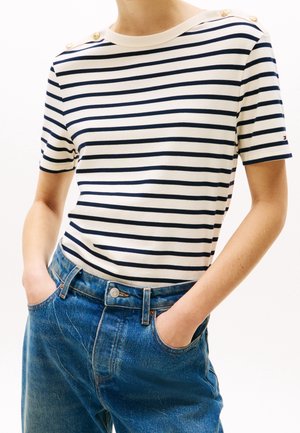Personne portant une chemise à manches courtes blanche et bleu marine rayée avec des boutons sur les épaules, rentrée dans un jean en denim bleu, les mains dans les poches.