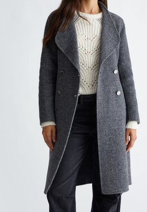 Femme vêtue d'un long manteau gris croisé sur un pull en tricot blanc et un pantalon noir, debout devant un fond uni.