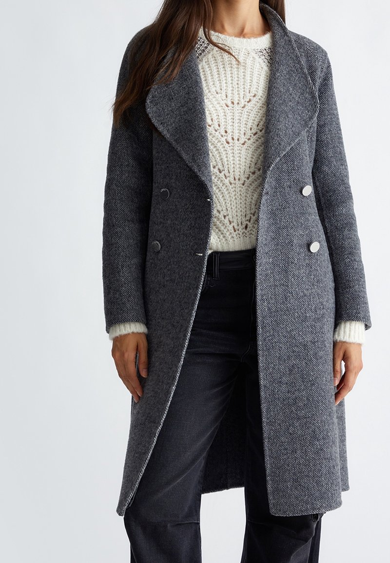 Femme vêtue d'un long manteau gris croisé sur un pull en tricot blanc et un pantalon noir, debout devant un fond uni.