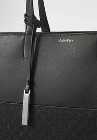 Černá taška přes rameno vyrobená z hladkých a strukturovaných materiálů, s kovovým štítkem ve stříbrné barvě, prošívanými detaily a logem na přední straně.