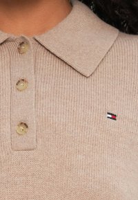 Beige kötött pólóing, gallérral és három gombbal. Az mellkason egy kis hímzett logó található piros, fehér és kék színekben.