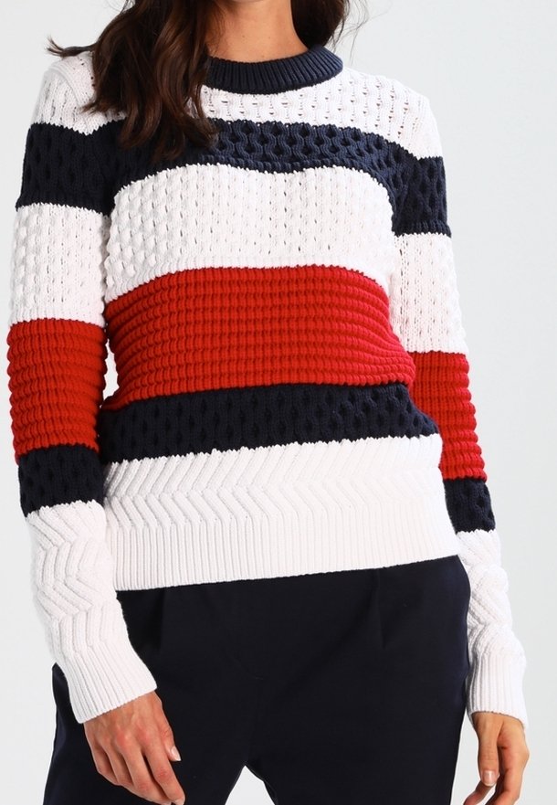 Kvinna som bär en stickad tröja med horisontella vita, marinblå och röda ränder, i kombination med mörka byxor.