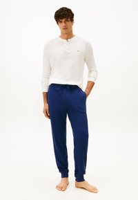 Maglietta henley bianca a maniche lunghe con un piccolo logo, abbinata a pantaloni del sudore blu con cintura regolabile e polsini alla caviglia. Vestibilità semplice e rilassata.