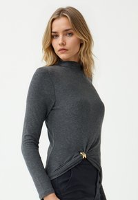 Blonde vrouw met golvend haar, gekleed in een aansluitend grijs shirt met lange mouwen dat op de taille is geknoopt, en een donkere broek, staand voor een effen achtergrond.