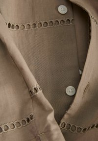 Hellbrauner Stoff mit Knopfverschluss weist dekorative Aussparungen entlang des Rands auf. Glatte Textur mit einem dezenten Glanz.