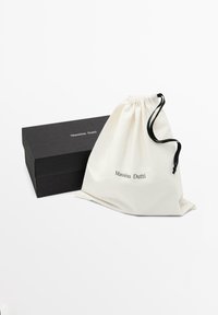 Sac à cordon en coton blanc cassé avec le logo "Massimo Dutti" imprimé, accompagné d'une boîte rectangulaire noire. Texture lisse, design minimaliste.