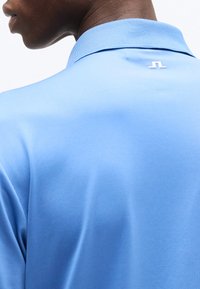 Άνδρας που φοράει ανοιχτό μπλε πόλο πουκάμισο με ριμπ ύφασμα στον γιακά και μικρό λευκό λογότυπο στο πάνω πίσω μέρος κάτω από τον γιακά.