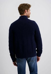 Dunkelblauer gestrickter Pullover mit hohem Kragen, gerippten Ärmeln und lockerer Passform, kombiniert mit hellblauen Jeans.