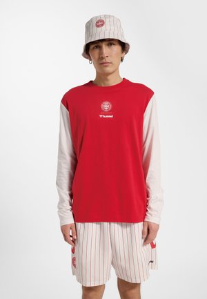 Junger Mann in roter und weißer Sportbekleidung mit DBU-Logos, darunter ein langärmeliges Shirt, gestreifte Shorts und ein passender Bucket Hat.