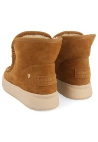 Las botas de tobillo de ante marrón presentan un forro suave, una forma redondeada y una suela de goma beige. La parte trasera incluye una marca y un sutil detalle texturizado.