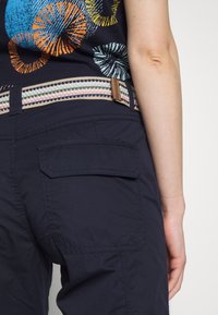 Gros plan d'une personne portant un pantalon bleu marine avec une poche rabat à l'arrière, une ceinture en tissu rayée, et un haut noir avec des motifs circulaires colorés.
