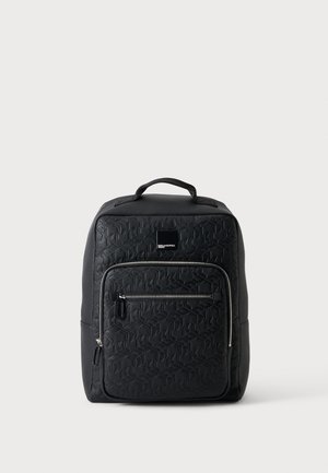 Sac à dos en cuir noir avec motif géométrique embossé, poche avant zippée, poignée supérieure et fermetures éclair argentées sur un fond uni clair.