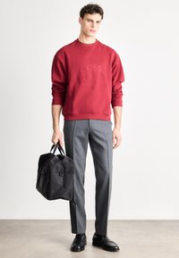 Κόκκινο φούτερ με ανάγλυφο λογότυπο "BOSS", γκρι ραμμένο παντελόνι, μαύρα δερμάτινα παπούτσια και μαύρη τσάντα duffel με διπλές λαβές.
