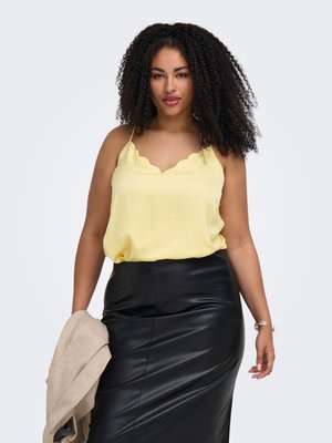 Femme aux cheveux bouclés portant un chemisier sans manches jaune et une jupe en cuir noire, tenant une veste beige dans une main.