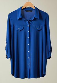 Camicia azzurra con collo, due tasche sul petto e maniche a 3/4. Realizzata in tessuto liscio con una leggera trama.