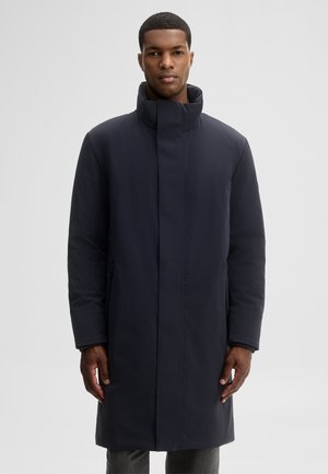Mann steht und trägt einen langen dunkelblauen Steppmantel mit hohem Kragen vor einem einfarbigen hellgrauen Hintergrund.