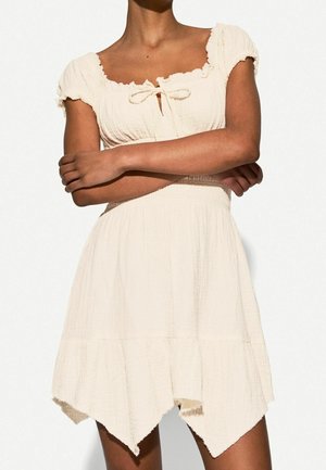 Mujer con un vestido corto crema con mangas abullonadas, escote cuadrado y detalle de lazo en el pecho, de pie con los brazos cruzados.