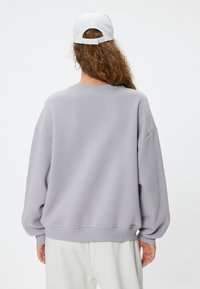 Sudadera oversized gris claro con hombros caídos y puños acanalados, con un escote redondo y una textura suave. Gorra blanca llevada al revés.