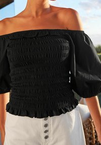 Černý top bez ramínek s smockovanou texturou, nabíranými rukávy a volánkovým lemem. Kombinováno s bílými kalhotami s vysokým pasem, které mají detaily s knoflíky.