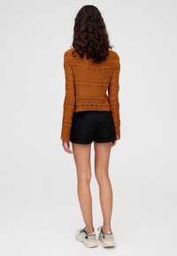 Cardigan all'uncinetto arancione con maniche lunghe, design con schiena scoperta, abbinato a pantaloni corti neri e sneakers chiare.
