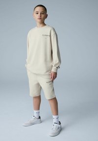 Ensemble sweat-shirt et short beige, en coton, coupe décontractée, poignets côtelés, détails du logo en brun foncé, assorti à des baskets blanches et des chaussettes.