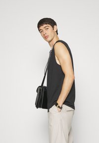 Maglietta nera senza maniche con scollatura rotonda, abbinata a pantaloni beige chiaro. Una borsa a tracolla nera presenta dettagli in metallo argentato e cuciture.