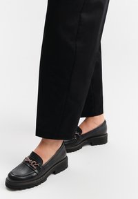 Czarne skórzane mokasyny o teksturowanym wykończeniu mają szeroką elastyczną taśmę i metalowy akcent. Gruba, solidna podeszwa zapewnia przyczepność.