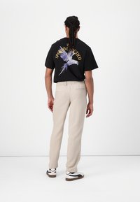 T-shirt noir avec un graphisme d'oiseau purple et du texte doré à l'arrière, associé à un pantalon beige décontracté et des baskets blanches avec des accents noirs.