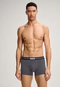 Modello maschile con un fisico in forma che indossa slip grigi con un elastico nero etichettato "JOOP!" in piedi contro uno sfondo chiaro e semplice.