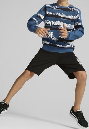 Felpa blu con motivi astratti bianchi, abbinata a pantaloni corti neri con logo e sneakers nere con suole bianche.