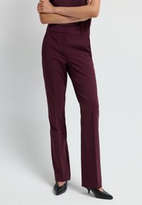 Pantalons ajustés bordeaux avec une coupe droite, fabriqués en tissu lisse. Comprend des passants de ceinture et une fermeture éclair. Associés à des talons noirs.