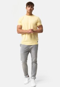 Gul bomulds t-shirt med rund hals, parret med lysegrå jeans og hvide sneakers. Modellen står med hænderne foldet foran sig.