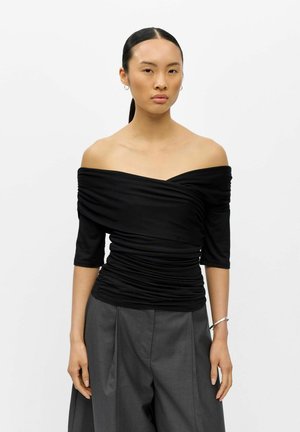 Kvinde med mørkt hår i en lav hestehale iført en sort off-shoulder rynket top og grå højtaljede bukser mod en ensfarvet hvid baggrund.