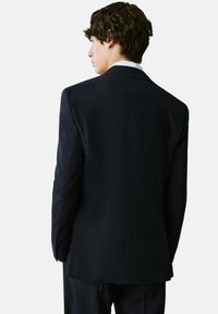 Mornarica blejzer s krojem koji ističe figuru, s rebrastim lapelima, jednim prorezom i teksturiranom tkaninom. Prikazan s leđa, detalji uključuju elegantnu siluetu.