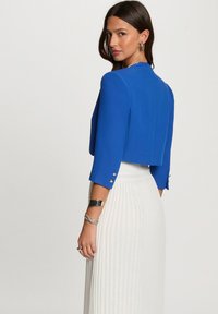Korte blauwe jas met driekwartmouwen en accenten van zilveren knopen, gecombineerd met een witte geplooide rok. Gladde stofstructuur.