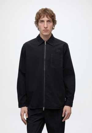 Junger Mann mit lockigem Haar, der eine schwarze Jacke mit Reißverschluss, einer Brusttasche und einem kleinen Logo vor weißem Hintergrund trägt.
