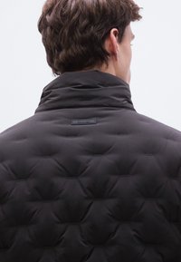 Μαύρο μπουφάν puffer με ψηλό γιακά, καπιτονέ υφή και μικρό λογότυπο στο πίσω μέρος. Λείο φινίρισμα με εφαρμοστή σιλουέτα.