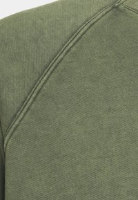 Tejido de sudadera de color verde oliva con una superficie texturizada y acanalada, y costuras visibles a lo largo de la costura. Diseño sencillo de cuello redondo.