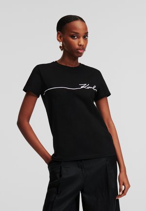 Zwarte katoenen t-shirt met korte mouwen, voorzien van witte cursieve tekst "Karl" op de borst. Soepele textuur, relaxed fit.