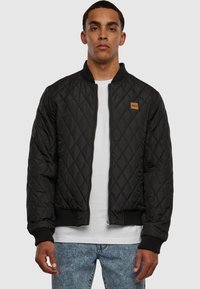 Čierna prešívaná bomber bunda s rebrovými manžetami a pásom. Má zips na prednej strane a malú hnedú nášivku na ľavej hrudi. Nosená cez biele tričko.