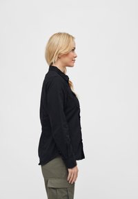 Camicia nera in cotone con bottoni indossata da una persona, caratterizzata da un colletto, maniche lunghe e un taglio dritto, abbinata a pantaloni cargo color oliva.