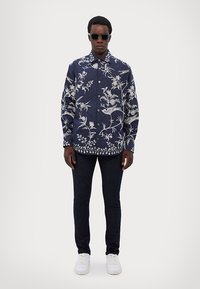 Camisa azul marino de botones con bordado floral blanco, combinada con jeans oscuros y zapatillas blancas. El diseño incluye mangas largas y cuello.